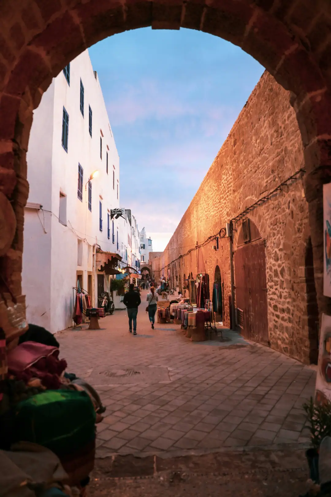 Gente paseando por una calle de la medina de Essaouira entre tiendas y una vieja muralla de piedra