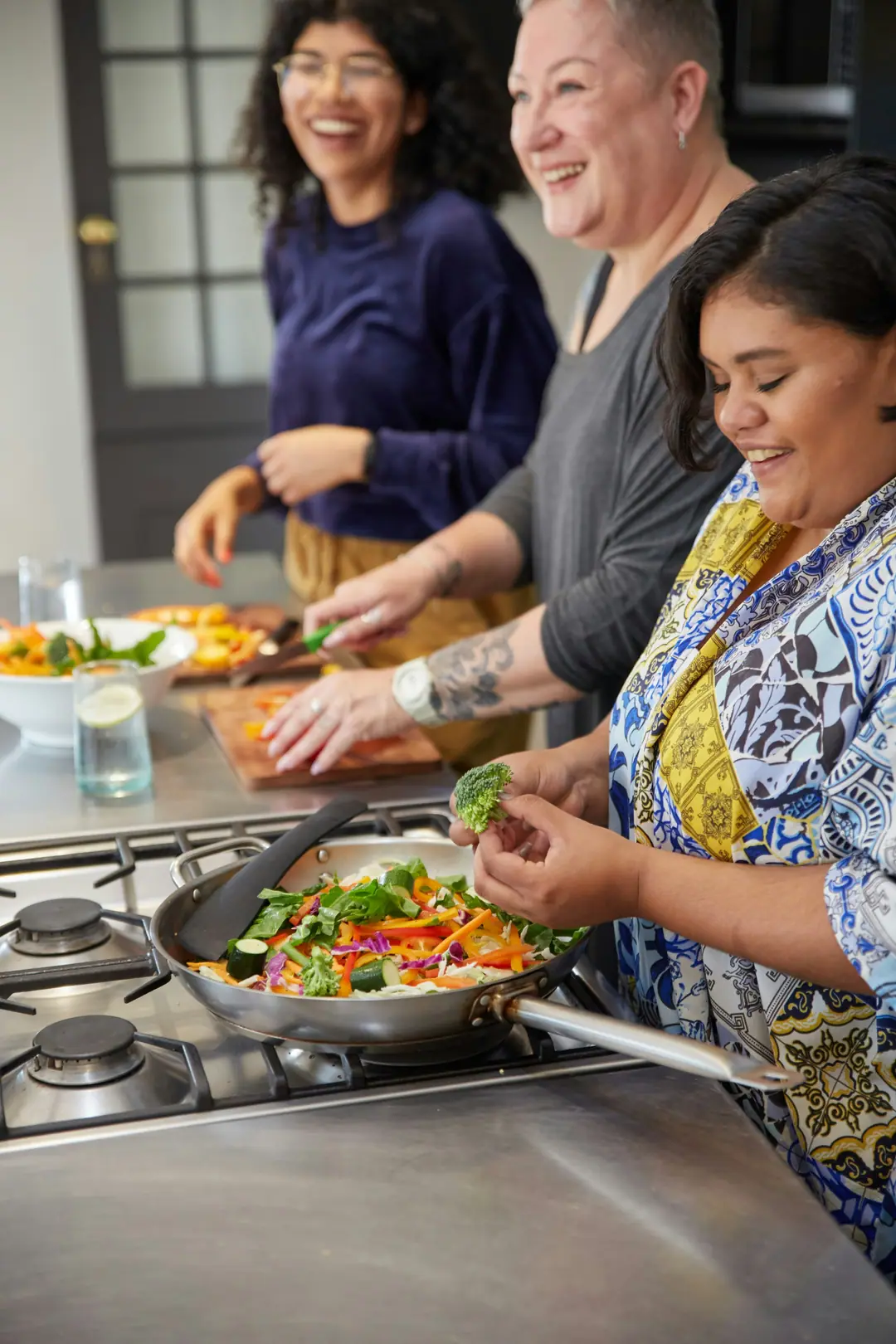 Three women laughing as they cook vegetables together at a stove