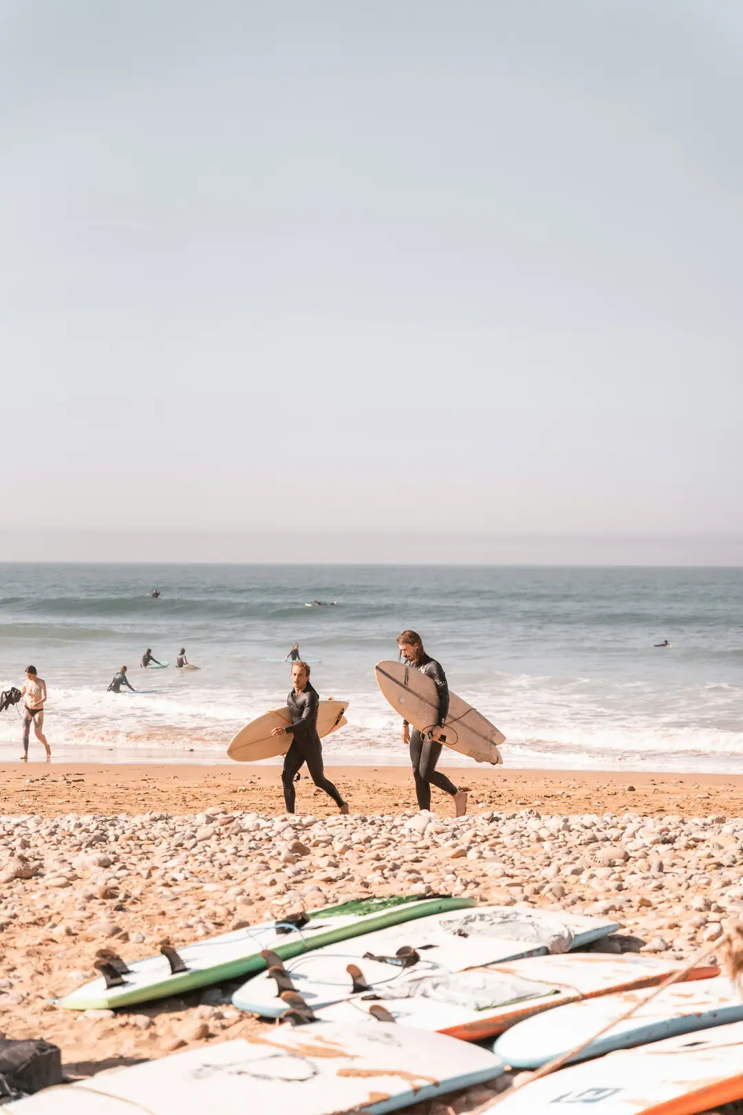 Two surfers carrying their boards to the waves past rows of surfboards on the sand